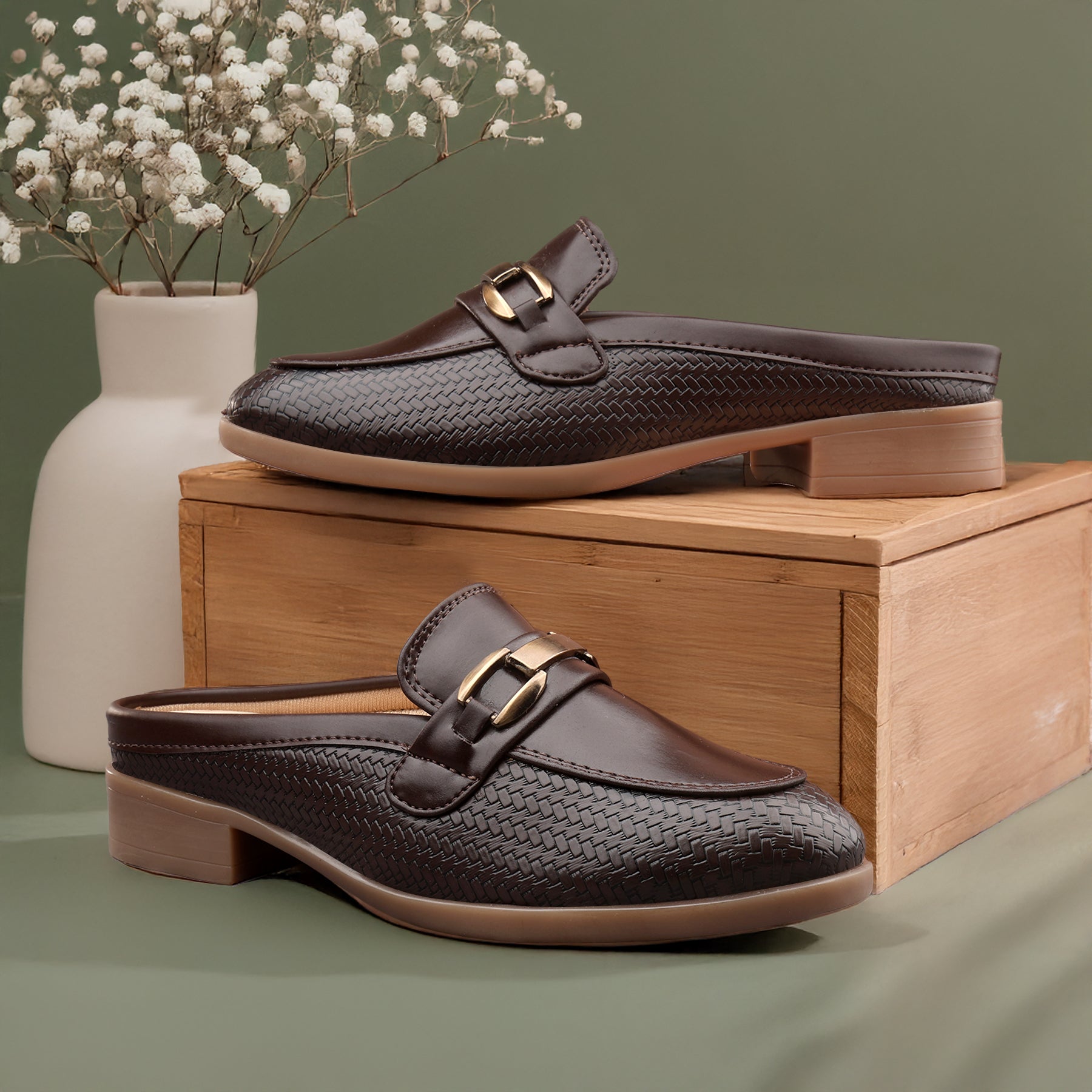 Brown woven loafers with gold buckles on a wooden box with a vase of white flowers in the background.