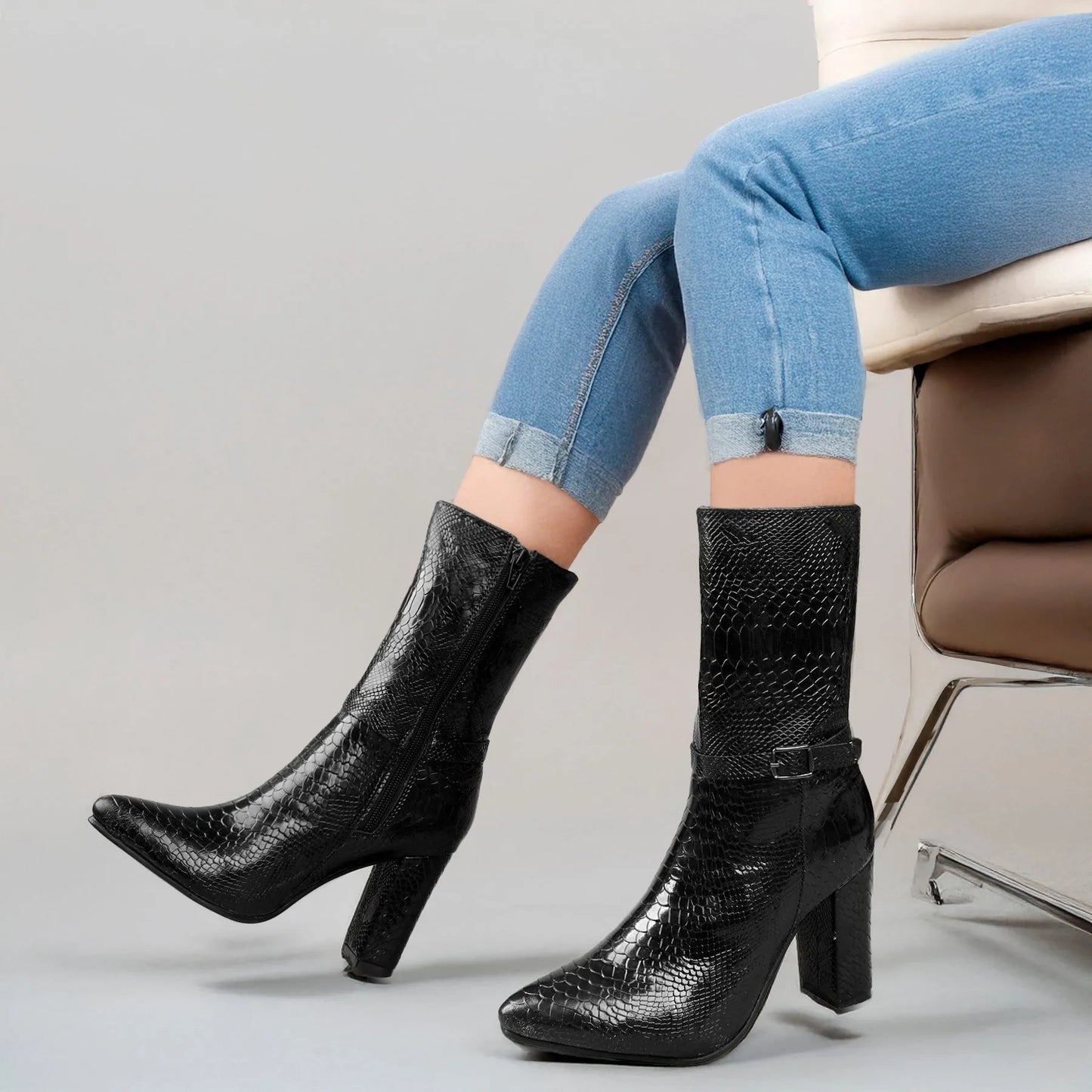 A pair of black snakeskin-textured ankle boots with block heels and side buckle details, shown on a model wearing light blue jeans seated on a chair.