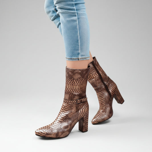 A person wearing brown snakeskin-patterned ankle boots with block heels and pointed toes, paired with light blue skinny jeans, standing against a warm beige studio background.