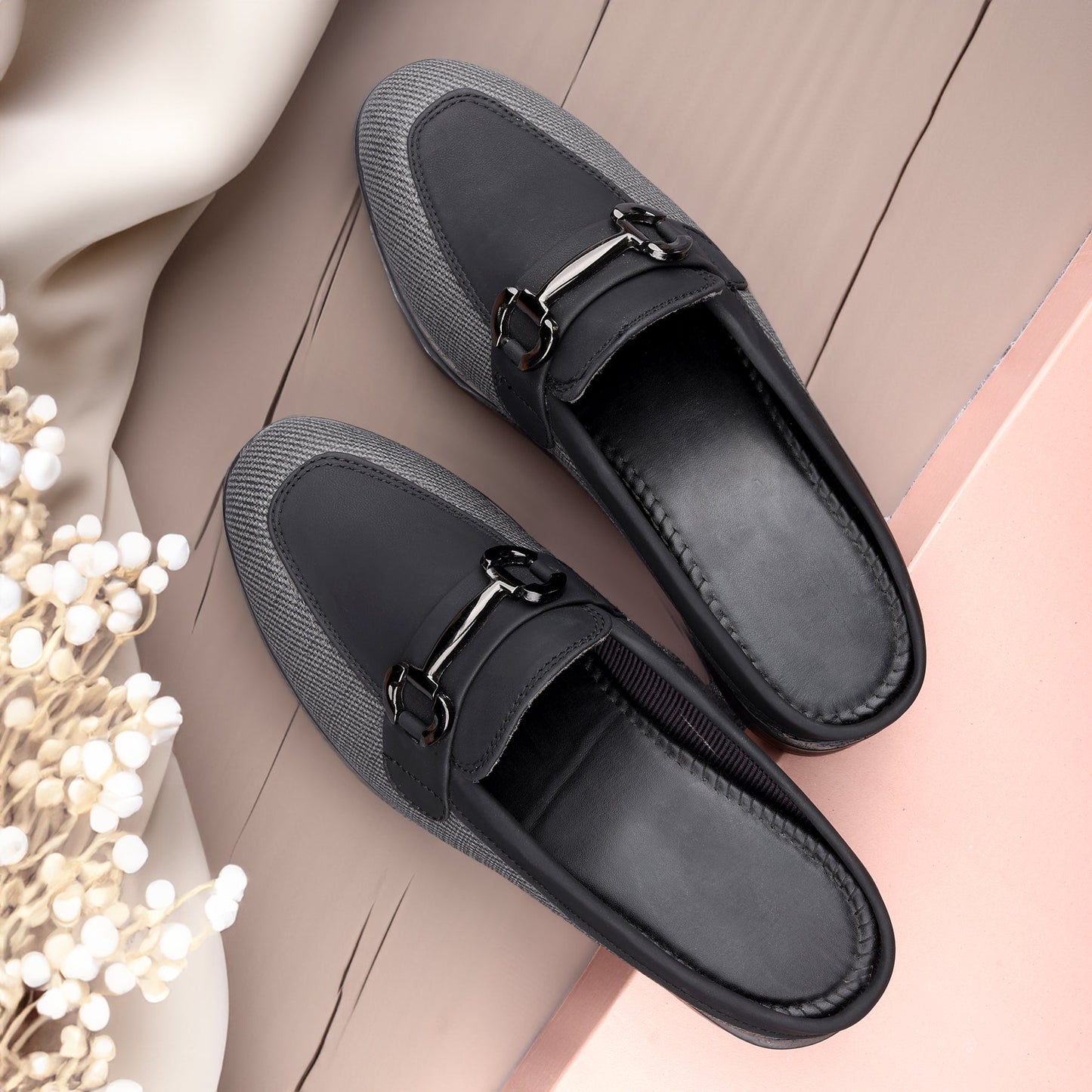 A pair of women's slip-on loafers in black and dark gray, styled on a beige wood plank surface with a soft pink block and white dried flowers as props.