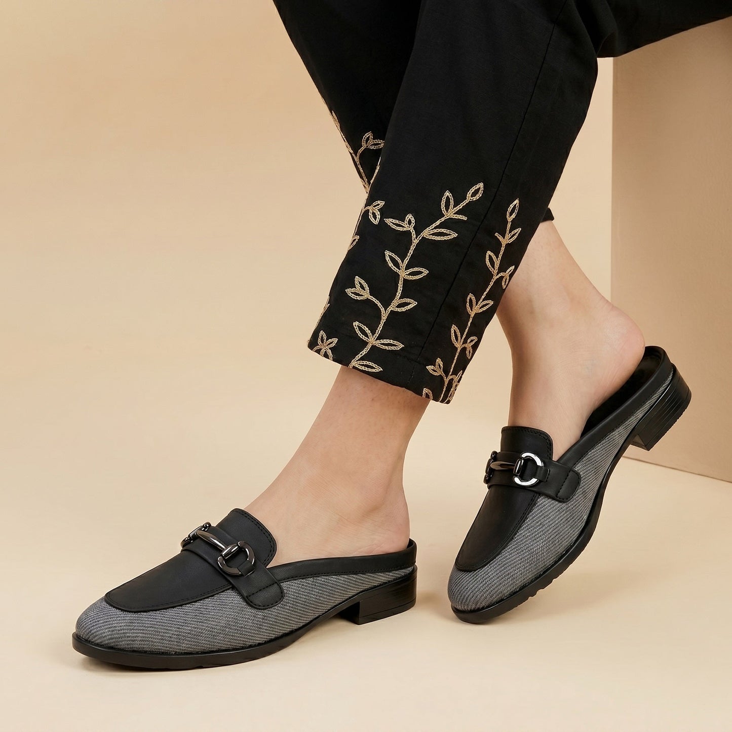 Black and gray loafers worn with black pants featuring embroidery on a beige background