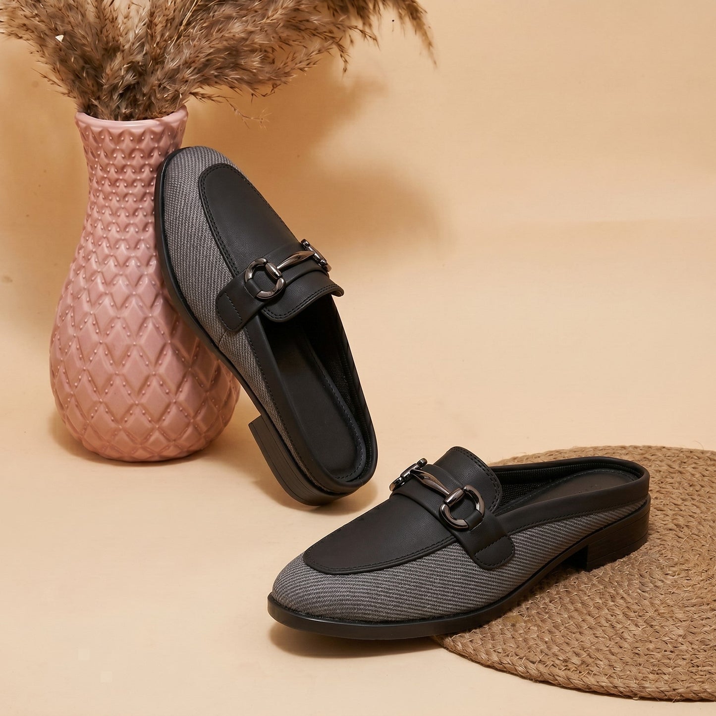 Pair of black loafers with a textured pattern on a beige surface with a pink woven vase and straw mat.