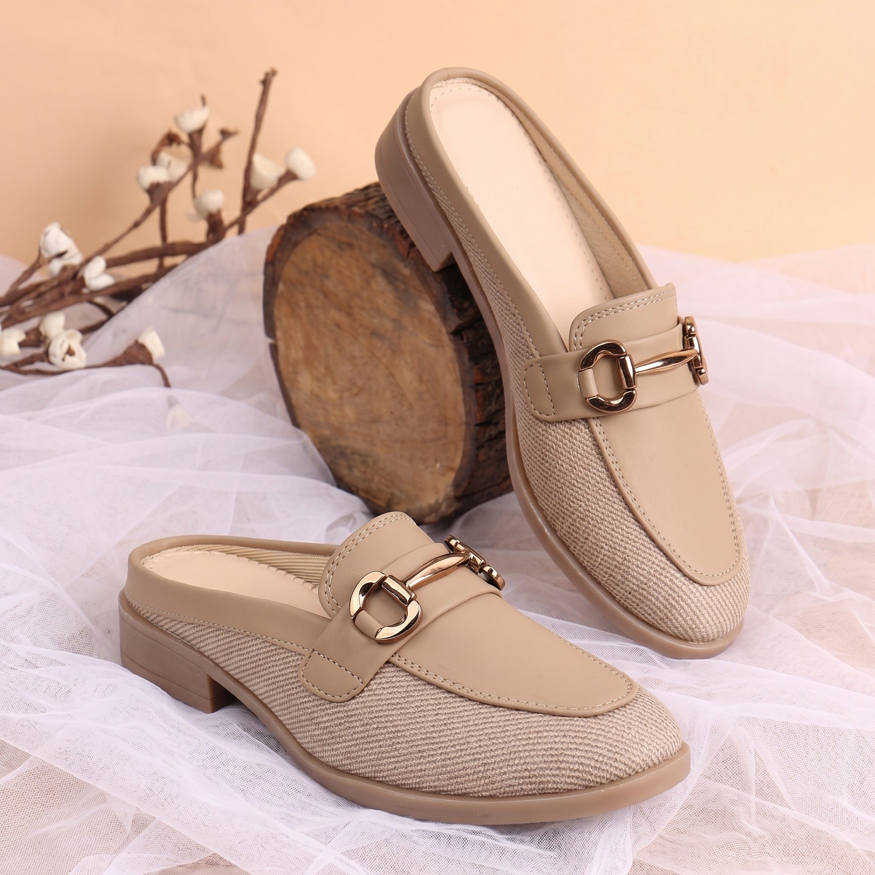 A pair of beige slip-on mule loafers with a textured woven fabric side, smooth leather toe cap, and a gold-tone horsebit detail, displayed on white tulle fabric with a rustic wood slice prop.