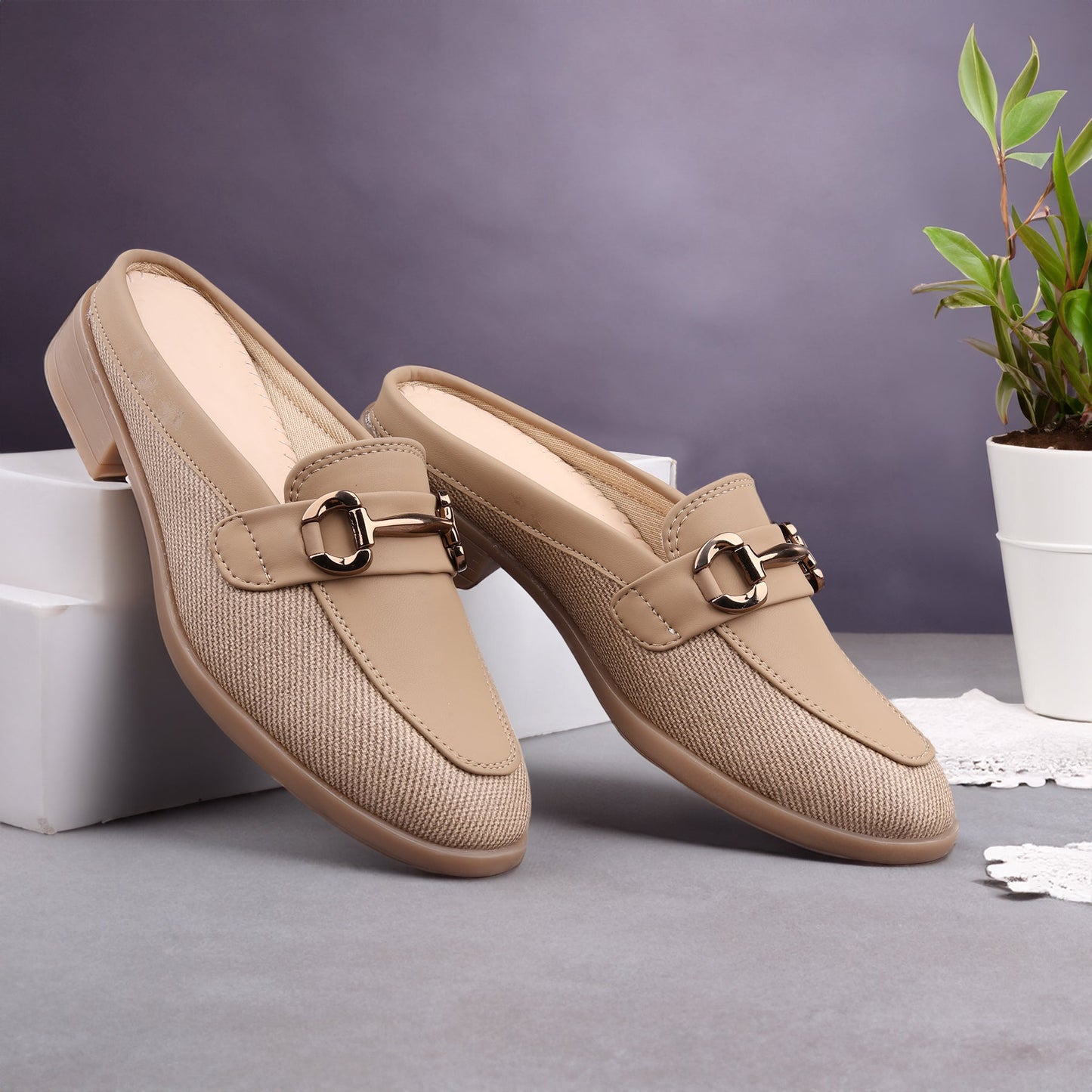 Pair of beige loafers with a gold buckle on a gray surface with a plant in the background.