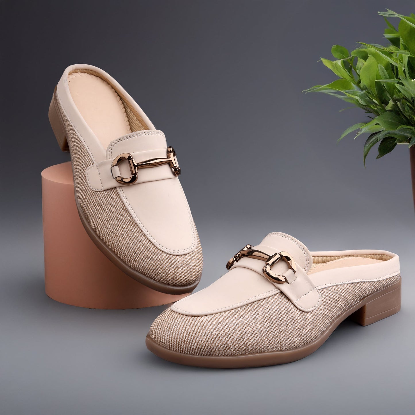 A pair of women's slip-on mule loafers in a light beige woven fabric with smooth beige leather trim and a gold-tone horsebit detail, displayed on a gray background with a pink block and a green plant.