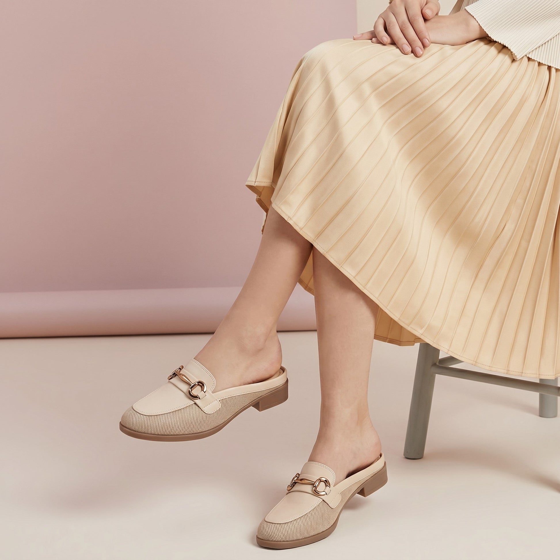 Person wearing beige loafers and a pleated skirt against a pink wall.