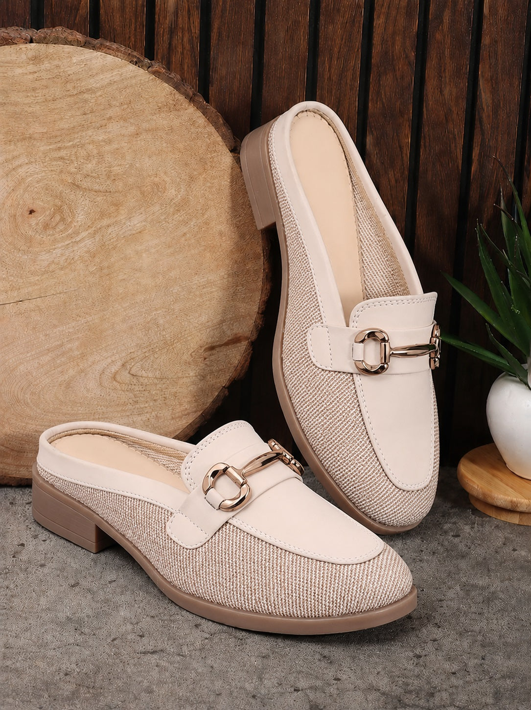 A pair of women's slip-on loafers with a beige and white textured woven upper, cream leather trim, and a gold-tone horsebit detail, displayed on a gray stone surface with a wood slice and a small plant in the background.