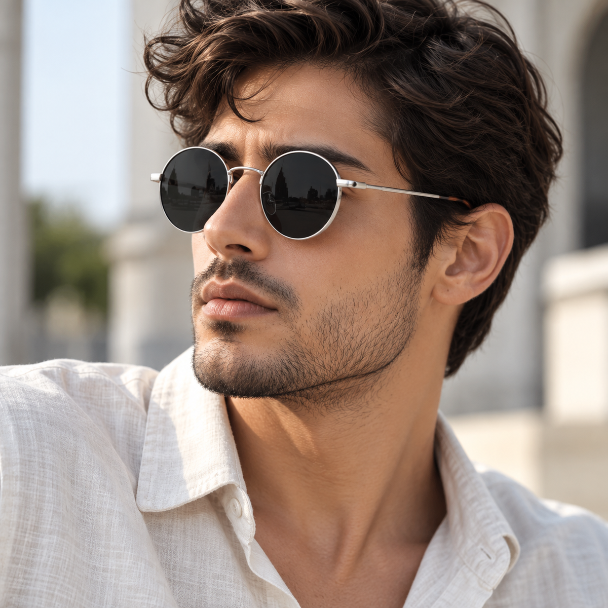 A close-up of a man wearing round silver-framed sunglasses with dark lenses and a light-colored linen shirt, set against a softly blurred outdoor architectural background.