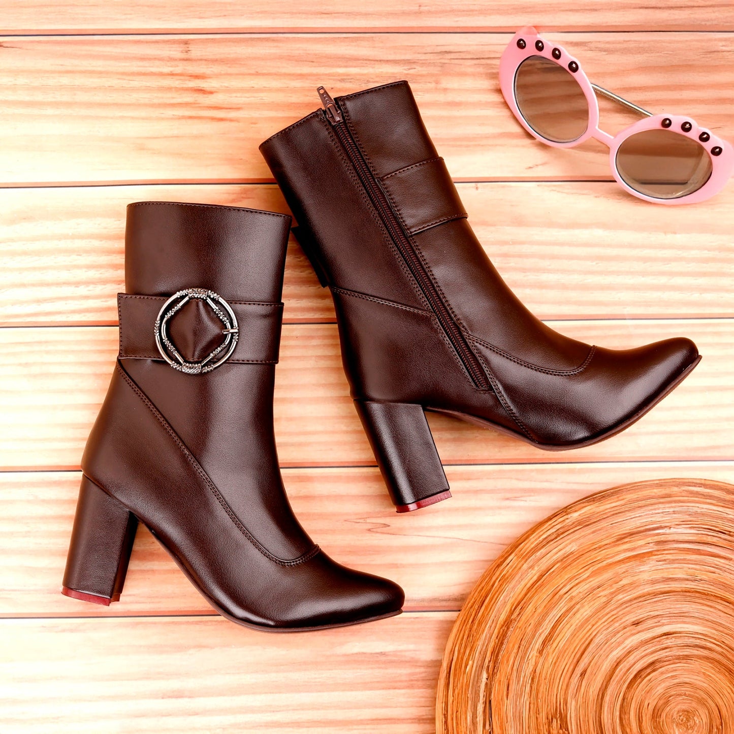 A pair of dark brown faux-leather ankle boots with block heels, displayed on a light wood background alongside pink sunglasses and a wooden tray.