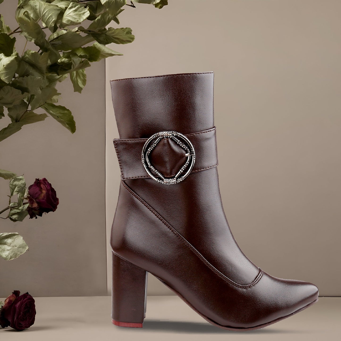 A dark brown faux-leather ankle boot with a block heel, pointed toe, and a decorative silver-tone buckle embellished with small clear rhinestones on the side, shown against a neutral beige background with dried roses and green leaves as props.