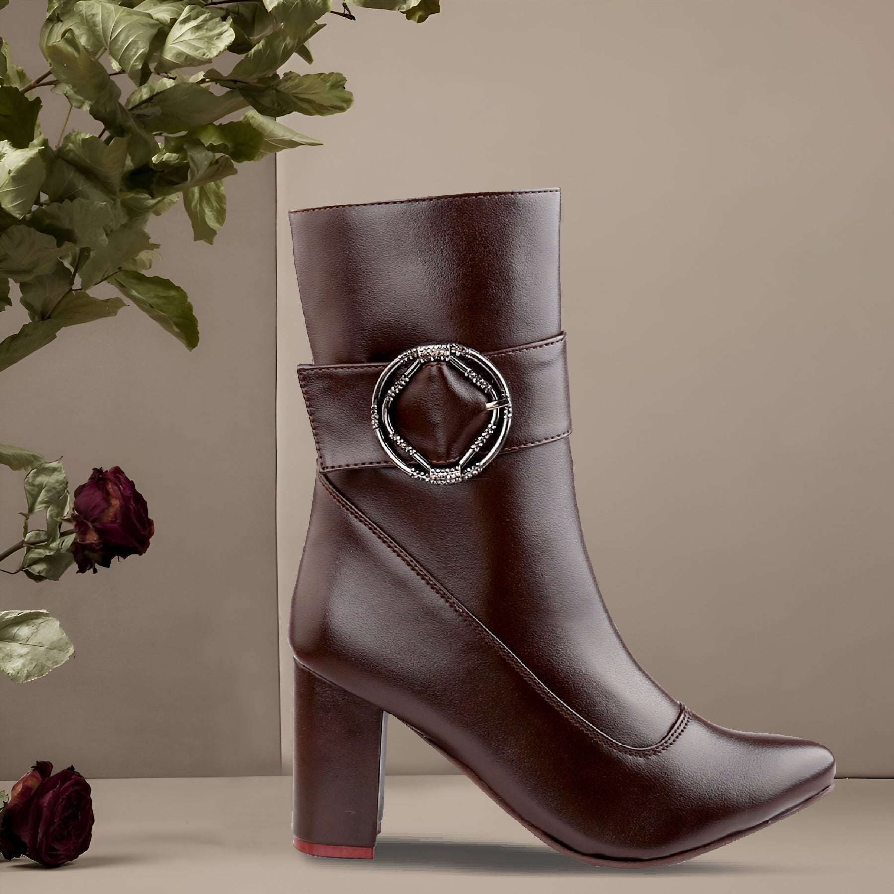 A dark brown faux-leather ankle boot with a block heel, pointed toe, and a decorative silver-tone buckle embellished with small clear rhinestones on the side, shown against a neutral beige background with dried roses and green leaves as props.