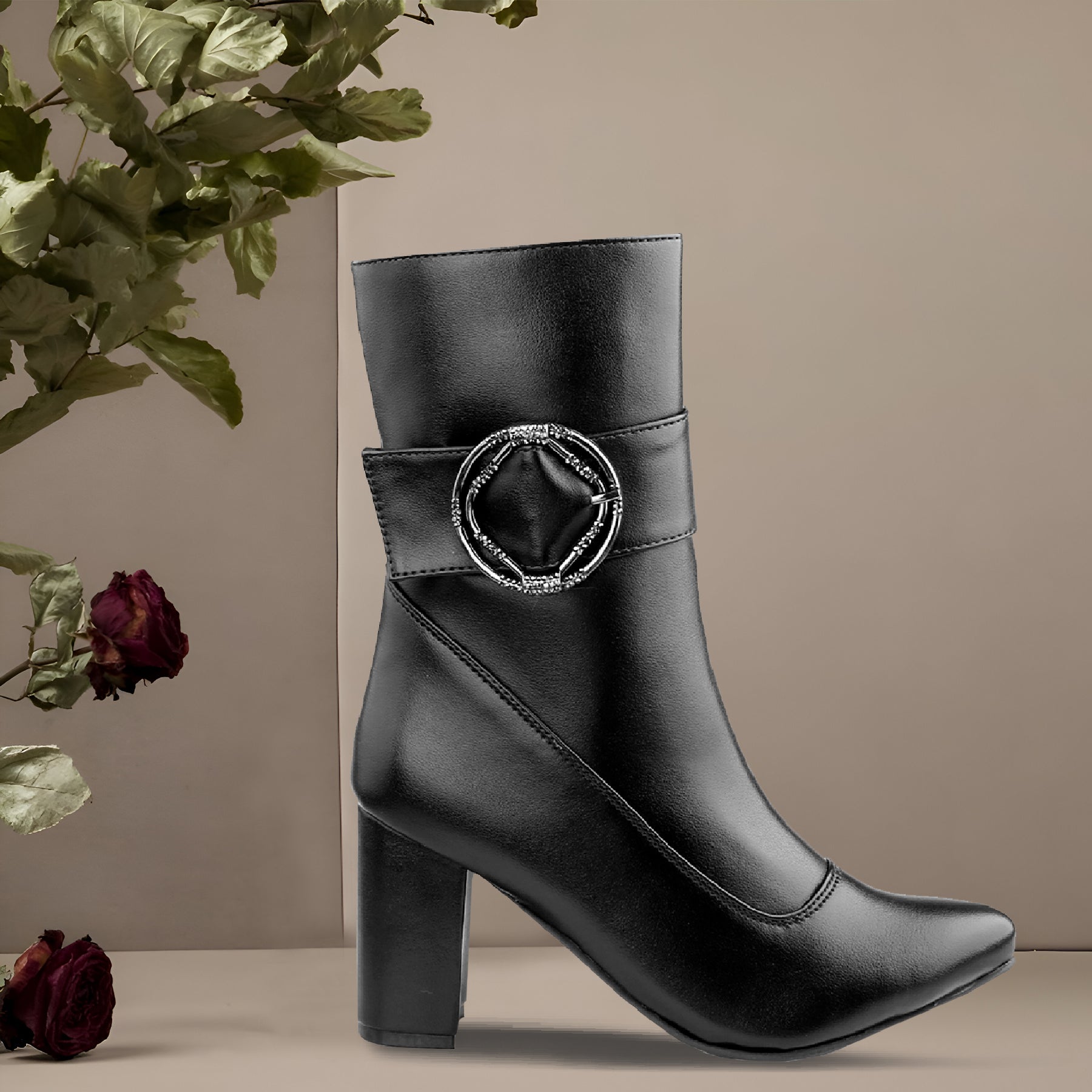 A black faux-leather ankle boot with a block heel and a silver-tone rhinestone-embellished geometric buckle, styled with dried roses on a beige background.