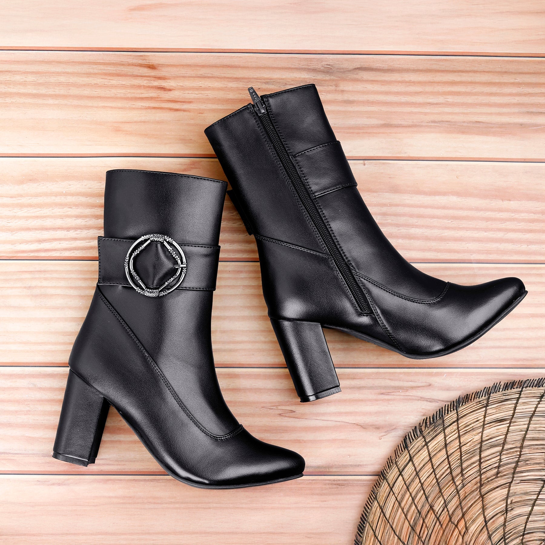 A pair of black leather ankle boots with block heels, displayed on a light wood plank background. One boot features a side zipper, and the other has a decorative silver-tone circular buckle detail on the side.