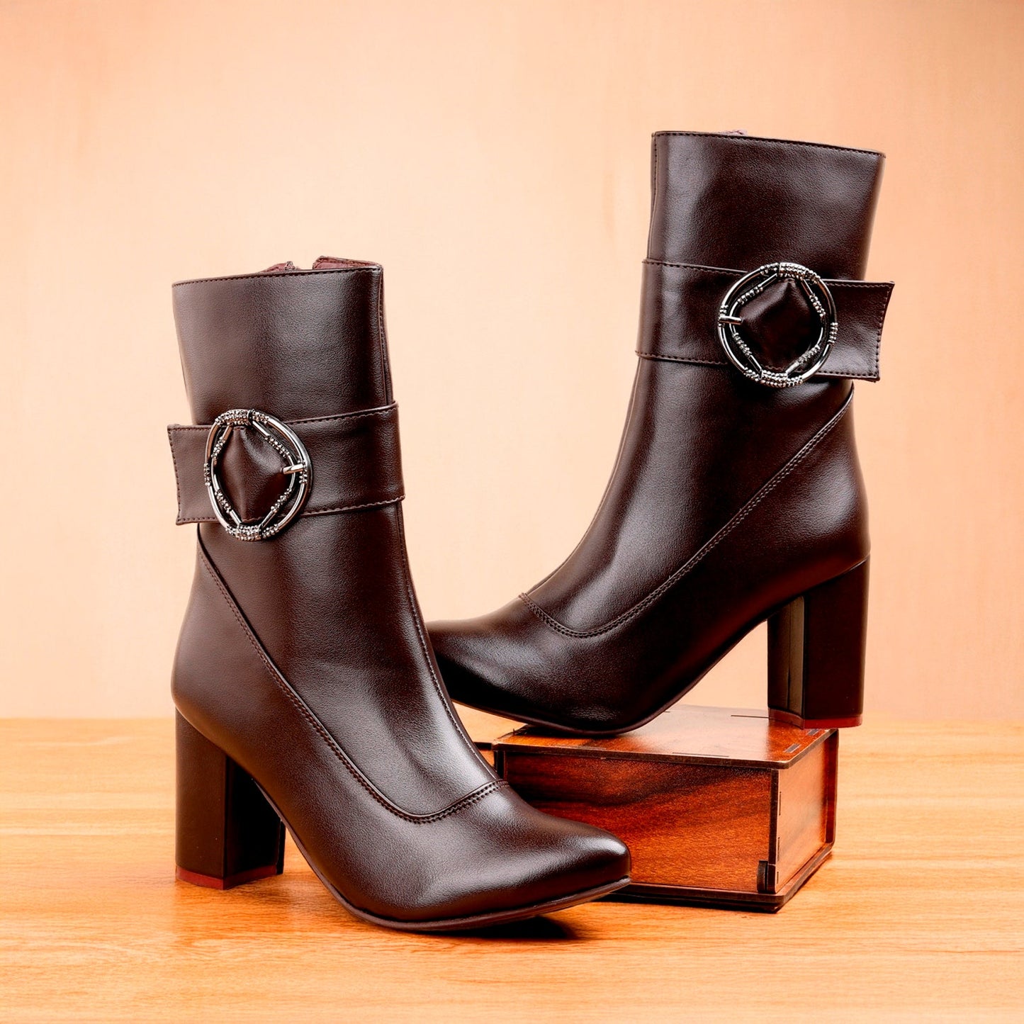 A pair of dark brown leather ankle boots with block heels and silver-tone buckle details, displayed on a wooden surface against a warm beige background.