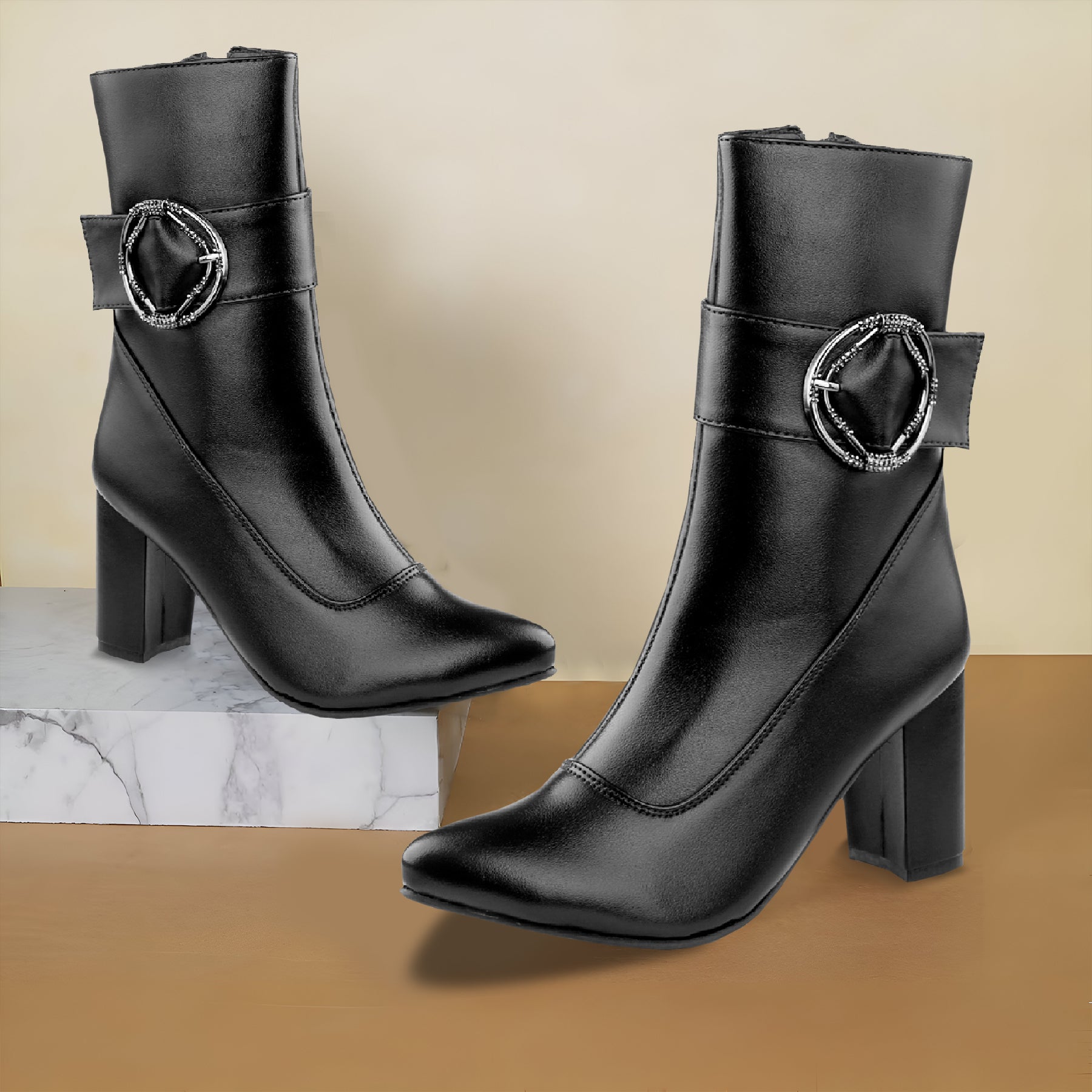 A pair of black leather ankle boots with block heels and a decorative silver-tone ring buckle strap on the side, displayed on a beige background with one boot standing on a marble block.