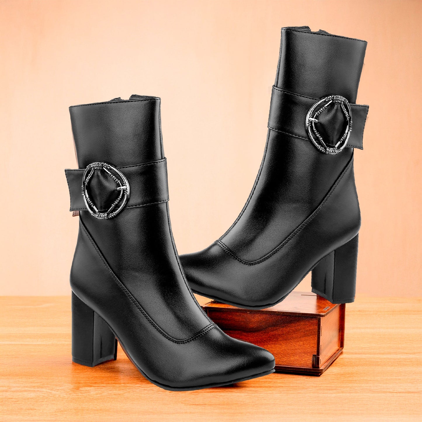 A pair of black leather ankle boots with block heels and a decorative silver-tone circular buckle detail on the side, displayed on a wooden surface with a wooden box prop against a warm peach background.