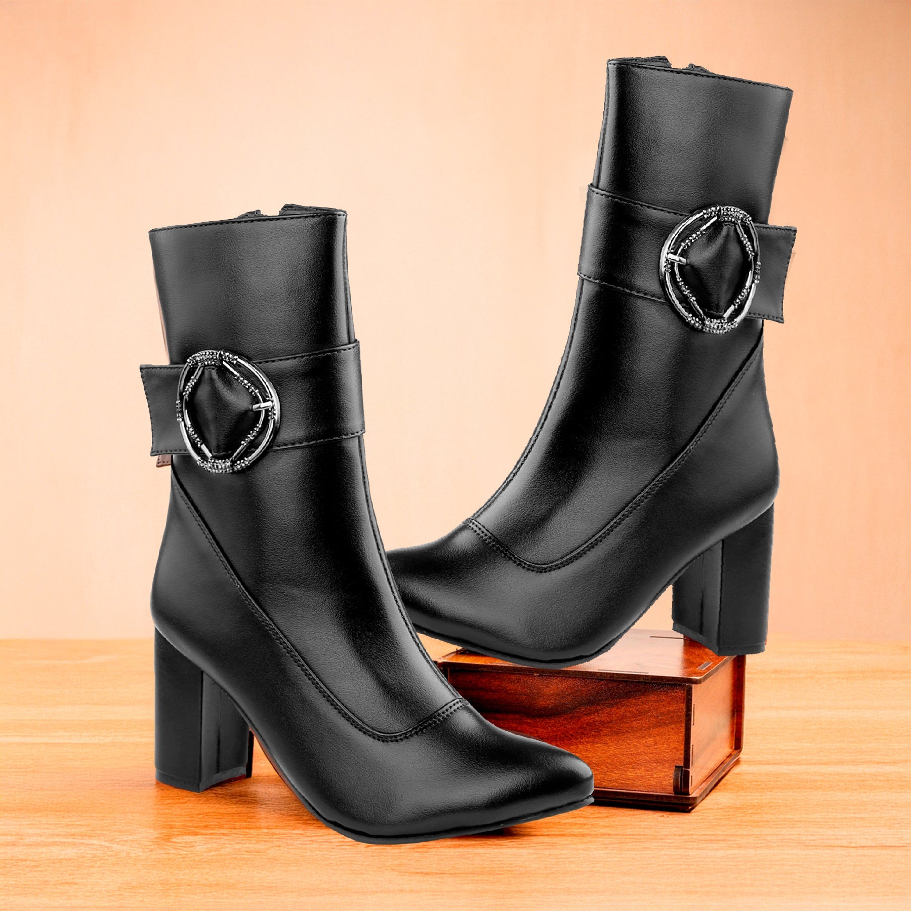 A pair of black leather ankle boots with block heels and a decorative silver-tone circular buckle detail on the side, displayed on a wooden surface with a wooden box prop against a warm peach background.