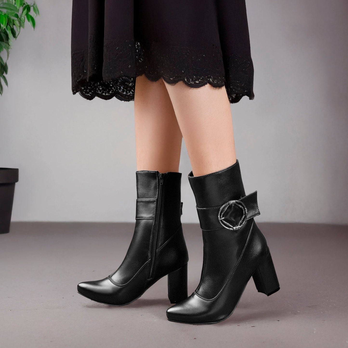 Black leather ankle boots with block heels and a decorative side buckle, shown on a model wearing a black skirt against a neutral gray background.