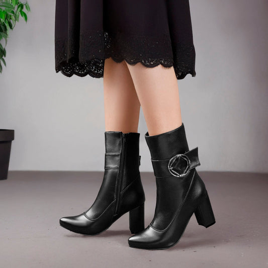 Black leather ankle boots with block heels and a decorative side buckle, shown on a model wearing a black skirt against a neutral gray background.