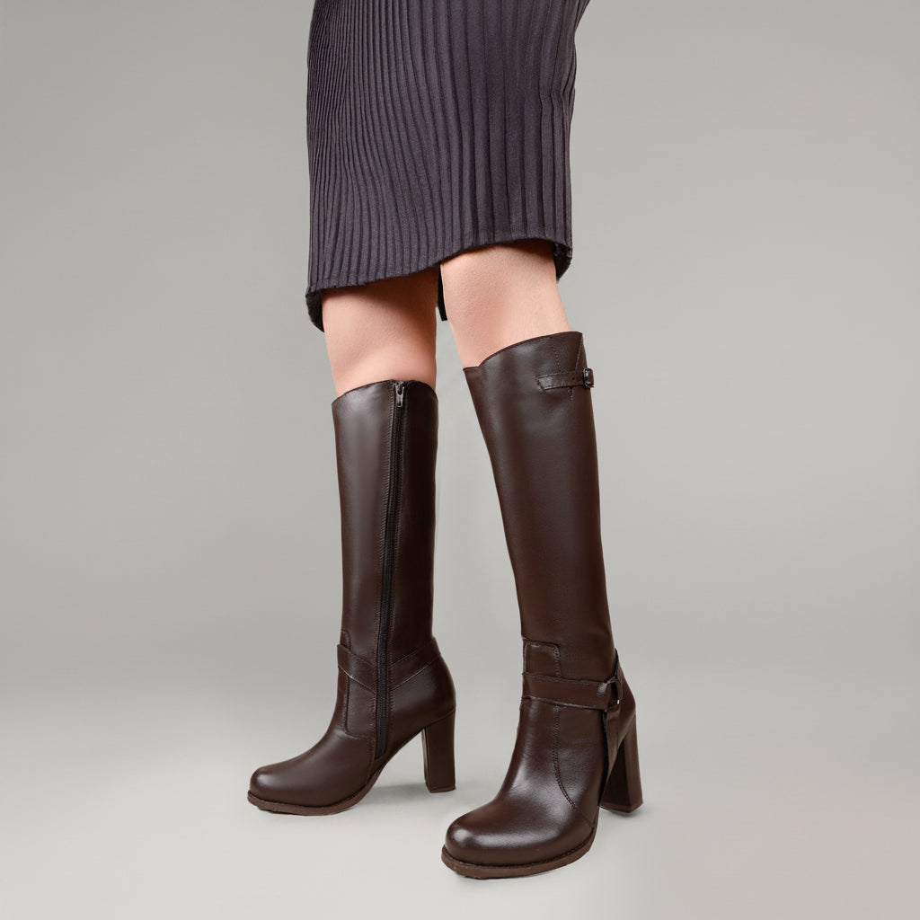 A person wearing dark brown knee-high leather boots with block heels and side zippers, paired with a dark ribbed skirt, against a solid warm beige studio background.