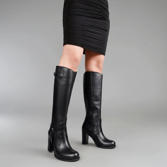 A person wearing a black mini skirt and black knee-high heeled boots with side zippers and buckle details, photographed against a plain gray studio background.