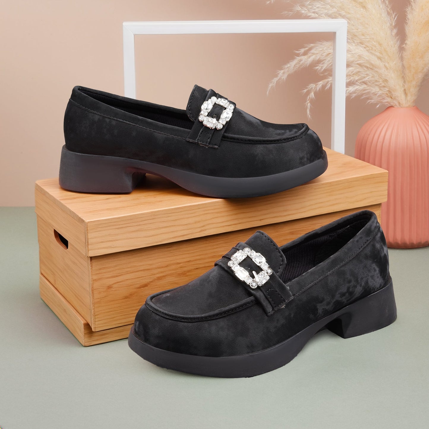 Black loafers with a decorative buckle on a wooden box against a beige wall.