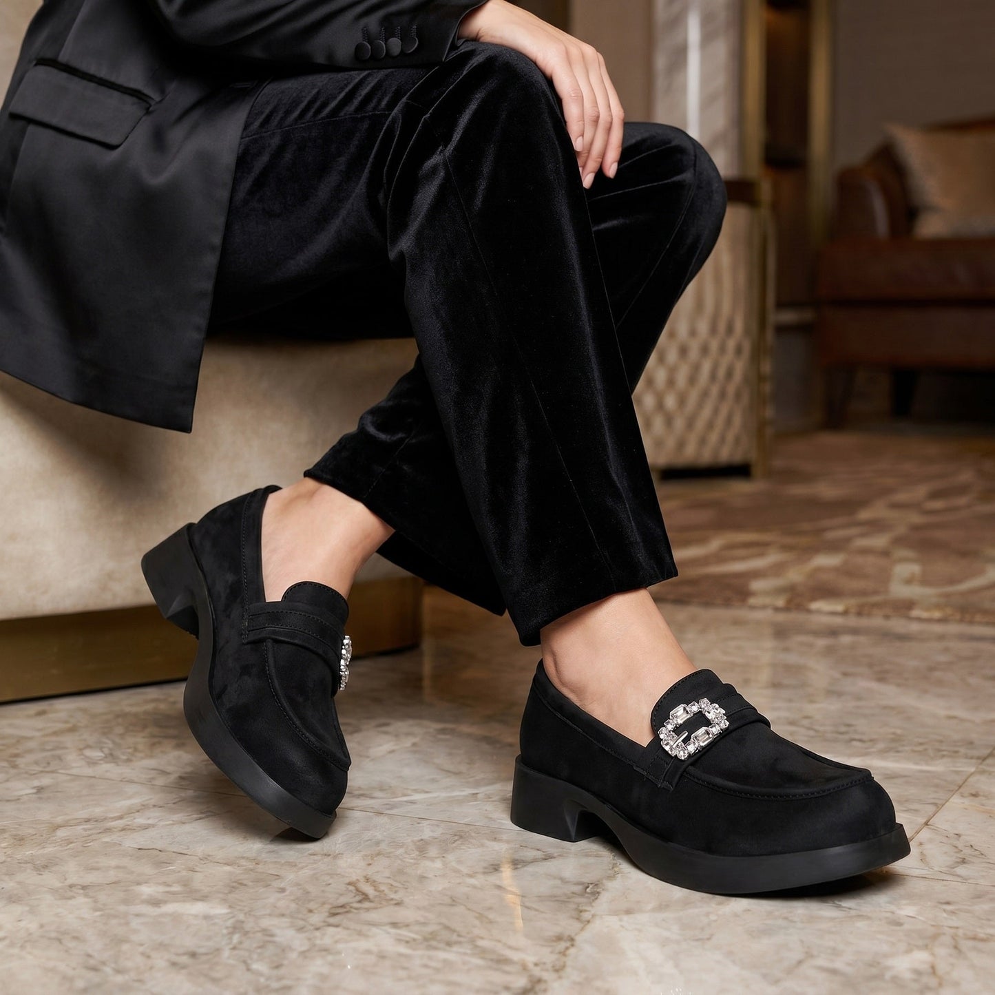 A person seated indoors wearing black velvet trousers and black loafers with a jeweled square buckle detail on the strap. The shoes are shown on a marble floor in a furnished room.