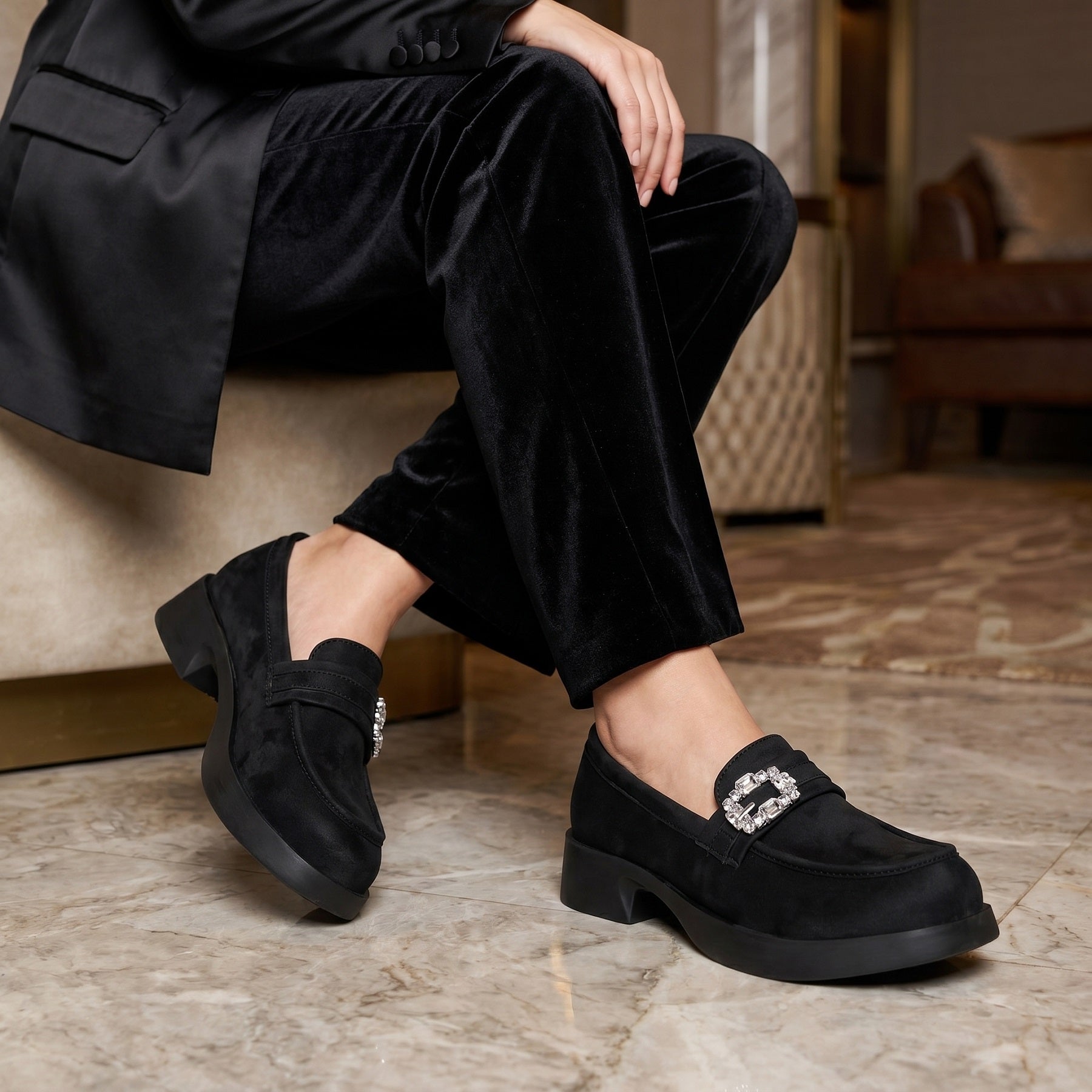 A person seated indoors wearing black velvet trousers and black loafers with a jeweled square buckle detail on the strap. The shoes are shown on a marble floor in a furnished room.
