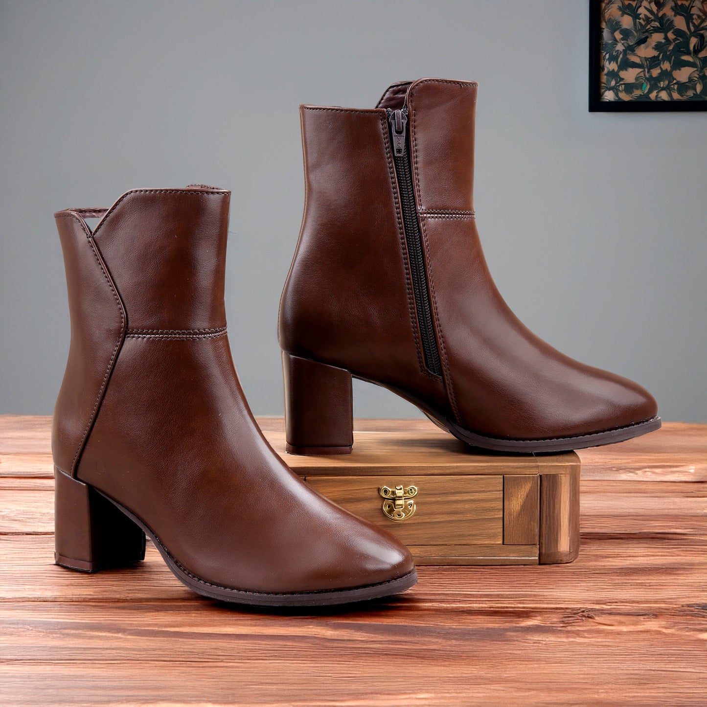 A pair of brown leather ankle boots with block heels and side zippers, displayed on a wooden surface with a wooden box in the background.