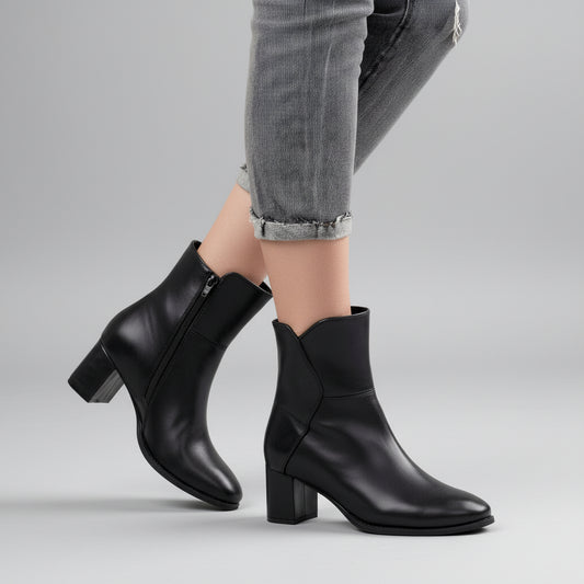 A person wearing black ankle boots with low block heels and pointed toes, paired with gray cuffed jeans, standing on a light wood floor.