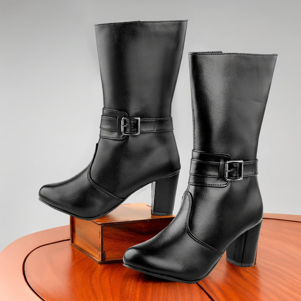 A pair of black leather mid-calf boots with block heels and ankle buckle straps, displayed on a wooden table against a light gray background.